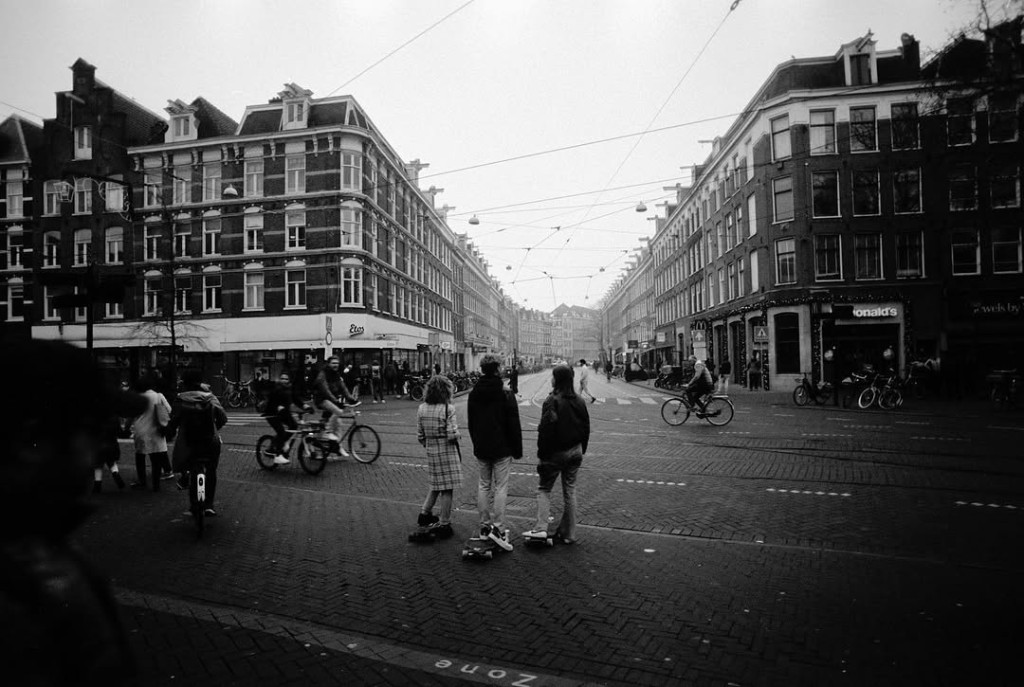 Black and white photograph of a cobblestone street intersection in Amsterdam, with cyclists and pedestrians under tram wires.